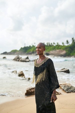 Flamboyanter schwuler schwarzer Mann im Luxuskleid posiert am malerischen Strand des Meeres. Transsexuelles, ethnisches Modemodel in langem, schickem Kleid und Accessoires blickt in eine Kamera. Meereswellen und Leuchtturm im Hintergrund.