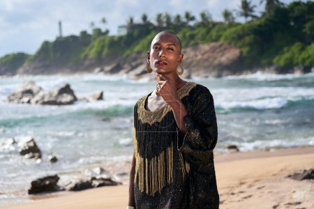 Outrageous gay black man in luxury gown poses on scenic ocean beach. Gender fluid ethnic fashion model in long posh dress and accessories looks at camera, touches face with a hand gracefully.