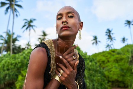 Trans sexual ethnic fashion model with brass jewelry accessories in elegant posture looks at camera. Epatage gay black man with golden rings, earrings, bracelets touches shoulder on scenic ocean beach