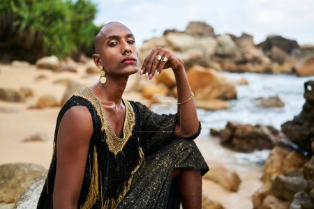 Androgynous ethnic fashion model in a luxury dress, jewelry sits on rocks by the ocean. Gay black person in jewellery and posh clothes poses gracefully in tropical seaside location portrait. Pride