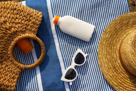 Overhead shot of summer essentials on striped towel with white sunscreen bottle, woven straw hats, stylish white sunglasses ready for sun-safe beach day by the ocean.