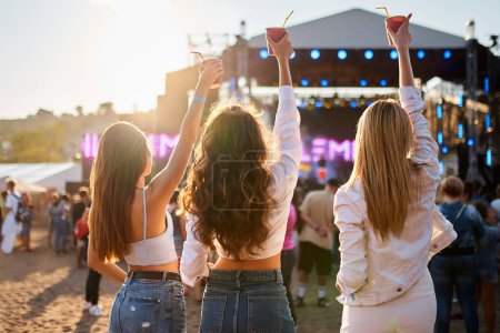 Casual summer outfits, sunset lighting, crowd and stage in background, girls celebrate together outdoors. Back view of three young women at beach music fest with raised drinks, enjoying concert vibes.
