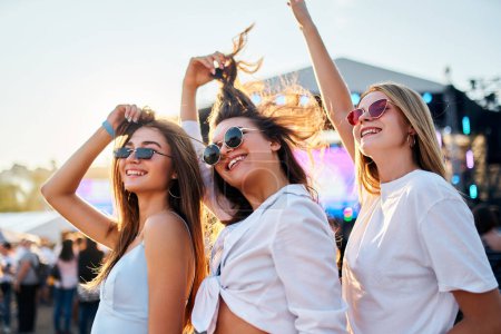 Friends dance, laugh with stage behind. Girls in sunglasses enjoy music at sunny beach fest. Casual summer wear, youth culture embraced at seaside concert. Holiday vibe as trio celebrates friendship.