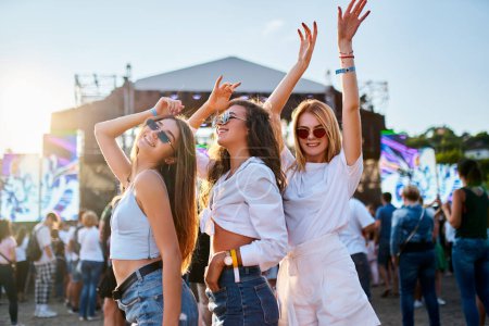 Sunlight illuminates the happy crowd, friends celebrate, party atmosphere by the ocean. Group of young women enjoying at a beach music festival, dancing together in summer outfits with sunglasses.