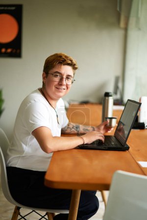 Pro engages in creative digital tasks, savoring tea, exemplifies inclusive workplace diversity. Transgender individual works at laptop in office space, sips hot beverage from steel thermos cup.