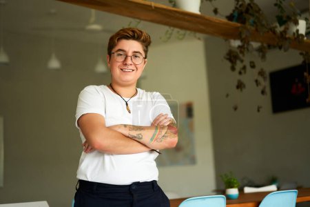 Inclusive workplace with natural light, plants. Confident Gen Z transgender person stands in modern office, arms crossed, smiling. Casual work attire, LGBTQ pride, authenticity at work reflected.