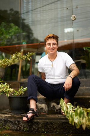 Contemporary digital nomad lifestyle, embracing workplace diversity, freedom. Smiling transgender individual works remotely outside plant-filled coworking space. Inclusivity in tech-focused.