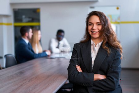 Colleagues discuss in background. Confident young businesswoman stands in modern office, smiling. Pro at work in formal attire, successful female leader, team meeting, workplace dynamics.
