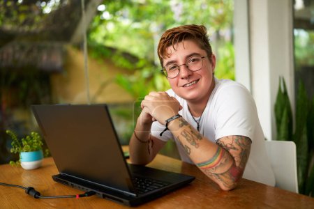 Inclusive work environment, tattoos visible, gender identity embraced, green plants background, youth culture. Transgender male person smiles at tropical cafe, laptop on table, earphones nearby.
