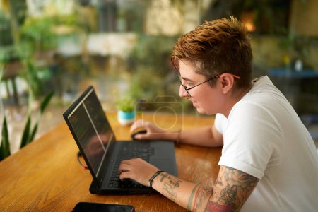 Focused on coding for mobile app, progressive workplace. Transgender software developer programs at wooden desk. Note tattoos, casual work attire. Green office signifies eco-friendly company culture.