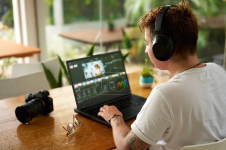Freelancer with tattoos wearing headphones edits film, digital camera on desk surrounded by plants by large window, daylight creative process. Professional video editor works on laptop in cozy cafe.