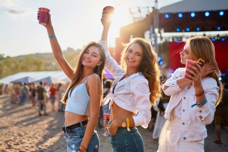Group of happy women celebrating at a beach music festival, sunlit scene with raised drinks, friends dancing, enjoying live concert outdoors, casual summer attire fun vacation atmosphere.