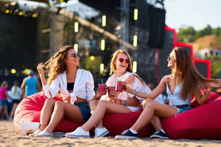 Fröhliche Freundinnen in lässiger Sommerkleidung genießen das Musikereignis, die Feier am Meer mit kalten Getränken. Gruppe fröhlicher Frauen räkelt sich auf roten Bohnensäcken und trinkt bei sonnigem Strandfest.