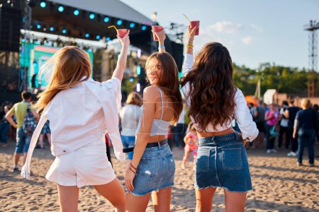 Freundinnen tanzen, genießen Sommerfest, Meereskulisse. Frauen feiern am sonnigen Strand mit Cocktails bei Live-Musik-Konzert. Lässige Mode, ausgelassene Stimmung unter den Festbesuchern.
