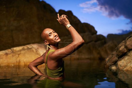 Photo for Smiling bipoc lgbtq model poses in water inside natural pool at night. Non-binary person shows jewelry - rings with gems on fingers, brass nose ring, golden earrings, bracelets, stands in a pond. - Royalty Free Image