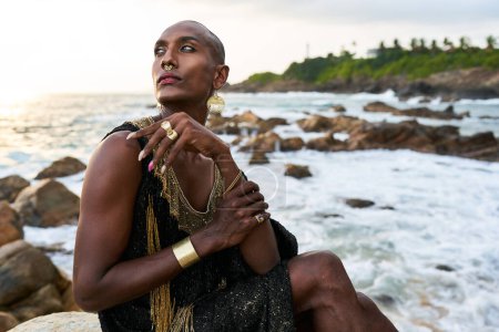 Queer schwarze Person im Luxuskleid, Schmuck sitzt auf Felsen in einem Ozean. Lgbtq ethnische Mode-Model trägt Schmuck in noblen Kleid gekleidet posiert anmutig in tropischen Küstenlage. Stolz auf LGBTQIA
