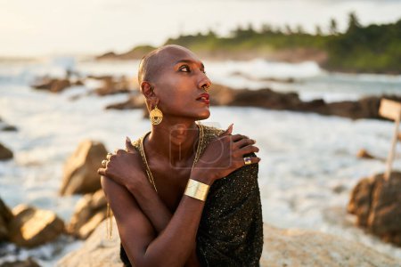 Photo for Non-binary black person in luxury dress, golden jewelry on beach rocks in ocean. Trans ethnic fashion model wearing jewellery in posh gown poses gracefully in tropical seaside location on a sunset. - Royalty Free Image