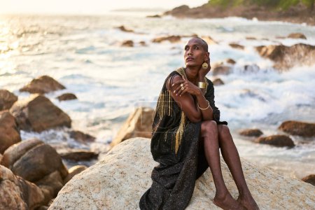 Non-binary bipoc fashion model in dress, brass jewelry sits on rocks by ocean. Trans sexual black person with rings, nose-ring, bracelets, earrings in posh clothes poses in tropical seaside location.