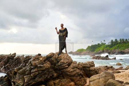 Geschlechtsneutrale schwarze Person posiert anmutig auf Felsen im Ozean. Androgynes Modell ethnischer Mode in schickem Kleid und Schmuck am felsigen Strand im Sturm. Stolzer Monat