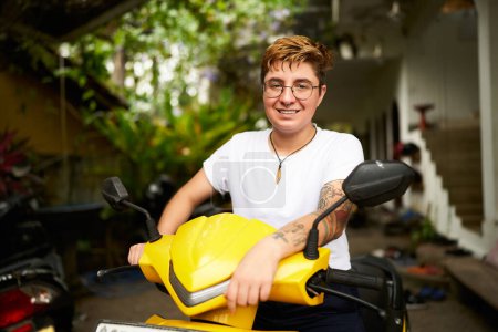 Short hair, casual style, holding handlebars ready for ride. Transgender person smiles, sitting on yellow scooter in tropical location. Scene reflects diversity, mobility, freedom in warm climate.