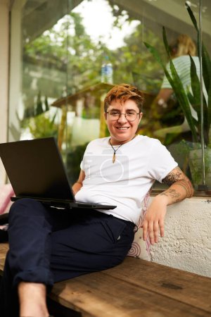 Relaxed attire, inclusive work environment evident. Transgender pro smiles while working on laptop in casual office space. Barefoot, shows comfort, tech engagement and confidence.