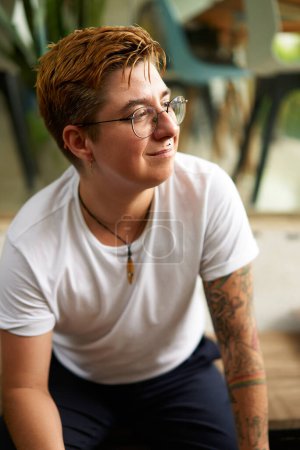 Short hair, tattoo embodies inclusion, empowerment in modern society. Positive transgender individual in casual setting, smiling confidently. Gender identity, workplace diversity.