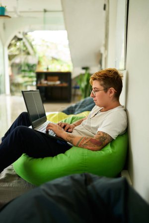 Espacio de coworking casual con luz natural, ambiente de trabajo amigable LGBTQ. Transgénero profesional trabaja de forma remota en el ordenador portátil, sentado en la bolsa de frijoles verdes. Confort, la productividad se fusiona en un entorno moderno.