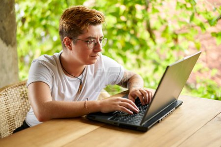 Non-binary entrepreneur focuses on remote work among green plants. Transgender digital nomad types on laptop at tropical coworking space. Inclusive, modern telecommuting lifestyle in natural setting.