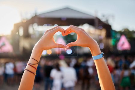Outdoor summer event, happy fans party, love symbol, festive vibe by the sea. Hands shape heart sign at sunset beach music fest, crowd enjoys live concert. Silhouette, sun glow, entertainment.