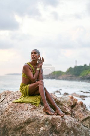Gender fluid black person poses gracefully sitting on high rock above ocean at sunset. Androgynous ethnic fashion model in dress on rocky hill by a storm at dusk. Pride month diversity concept.