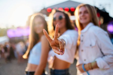 Smiling girls in trendy outfits dance, laugh, show peace sign. Group of female friends enjoy summer music festival on beach. Partygoers celebrate, have fun in sunset light at seaside concert event.