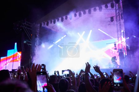 Fans record live performance on smartphones. Backlit stage with vibrant lights at outdoor music festival. Excitement in air, band plays hit songs. Summer night event, audience enjoys concert vibe.
