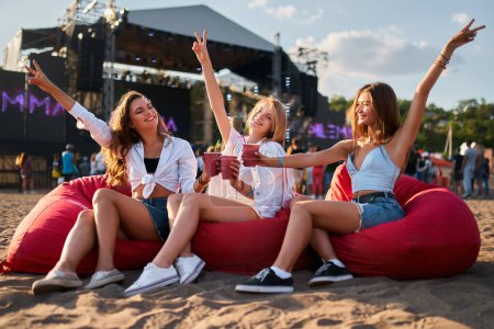 Women in casual summer wear, chilling, having fun, with stage in background. Group of joyful girls sit on red bean bags, enjoy cold drinks at sunny beach music fest. Carefree youth lifestyle scene.