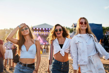 Drei Freunde tanzen, lachen in der Nähe der Bühne mit hellen Lichtern. Mädchen genießen das sommerliche Musikfestival am Sandstrand bei Sonnenuntergang. Modische junge Frauen in lässigen Outfits feiern bei Open-Air-Konzert.