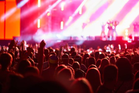 Fans wave hands, dance, celebrate. Concertgoers enjoy live music at outdoor fest, vibrant stage lights illuminate performers. Summer event vibe, festival season, night entertainment atmosphere.