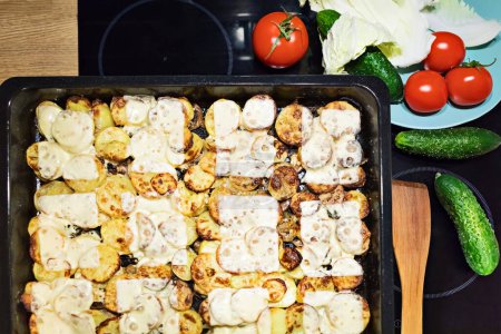 Téléchargez les photos : Casserole traditionnelle de pommes de terre maison, coupée en plastique, cuite au four avec du fromage jusqu'à ce qu'elle soit dorée, entourée de légumes (tomates, chou, concombres).. - en image libre de droit
