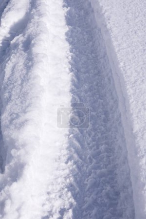 Téléchargez les photos : Le chemin d'empreintes sur une neige blanche étincelante du soleil, une chute de neige soudaine, la texture d'un fond neigeux sur la route avec des traces d'une roue de voiture, un climat givré, gros plan, verticalement - en image libre de droit
