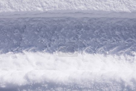 Téléchargez les photos : Le chemin d'empreintes sur une neige blanche étincelante du soleil, une chute de neige soudaine, la texture d'un fond neigeux sur la route avec des traces d'une roue de voiture, un climat givré, gros plan, horizontal - en image libre de droit