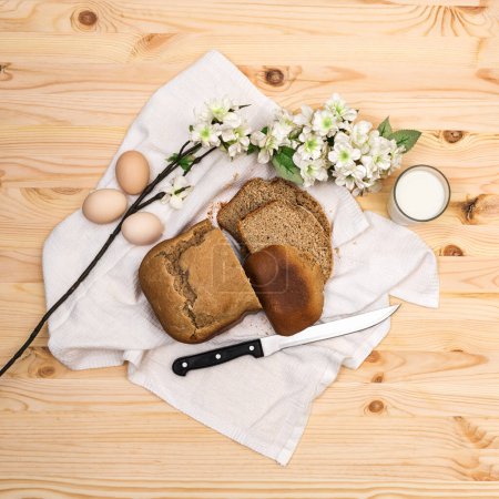 Téléchargez les photos : Un pain de seigle maison fraîchement cuit coupé avec un couteau sur une serviette blanche, un verre de lait et trois œufs de poulet sur une table en bois, nourriture simple et saine, carré, vue sur le dessus, plat - en image libre de droit