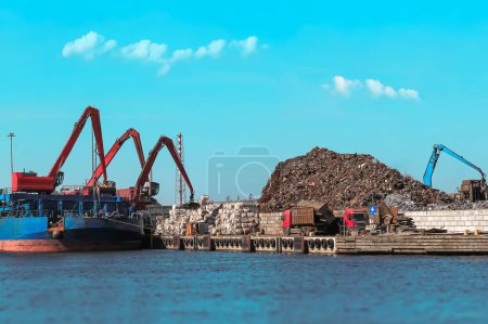 A large cargo ship is docked at a busy scrapyard. A massive crane is unloading scrap metal into a huge pile. Various industrial operations are in progress at the site.