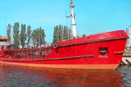 Ein großes rotes Frachtschiff legte zum Verladen im Seehafen an, umgeben von anderen Schiffen. Bäume und Gebäude im Hintergrund. Verwendung für den Transport von Gütern und Materialien.