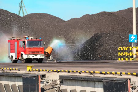 Un camion de pompiers rouge vif pulvérise de l'eau sur un grand tas de charbon noir, créant ainsi un bel arc-en-ciel, mettant en valeur la puissance des interventions d'urgence et des machines industrielles..