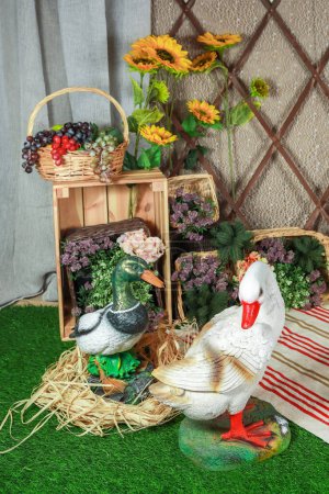A duck and goose couple stand beside a basket filled with fruit and flowers. The background is a rustic wooden fence with a sheer curtain on one side and a green grass floor.