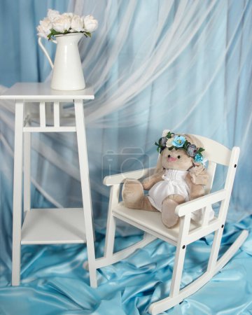 The rocking chair is made of wood and has a white finish. The stuffed bunny is white and has a floral headband on. The vase is filled with white flowers. The background is a light blue cloth.