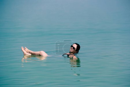 A woman in a black swimsuit floats in the buoyant Dead Sea water. The blue-green water and hazy sky create a serene scene. The high mineral content is known for its therapeutic properties.
