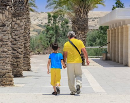 The father is wearing a yellow shirt and the son is wearing a blue shirt. They are both wearing  sandals. The father has his hand on the sons shoulder. They are walking towards a building.