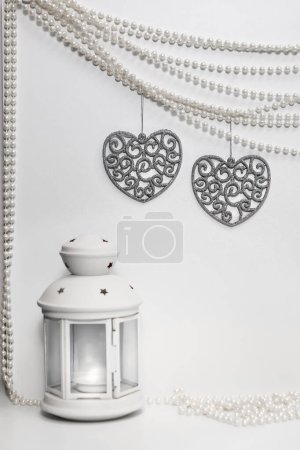 Two silver glitter hearts with flourishes hang from strings of white pearls against a solid white background with a white shabby chic lantern with a lit candle sitting in front on the white surface.