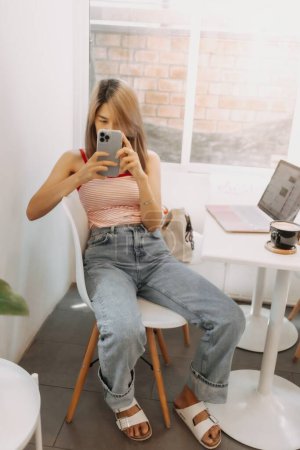 Cute freelance asian girl is using smartphone while working on laptop.