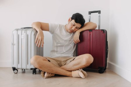Traurig enttäuschter asiatischer Mann sitzt mit seinem Gepäck da sein Reiseplan gestrichen wurde.