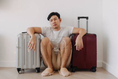 Traurig enttäuschter asiatischer Mann sitzt mit seinem Gepäck da sein Reiseplan gestrichen wurde.